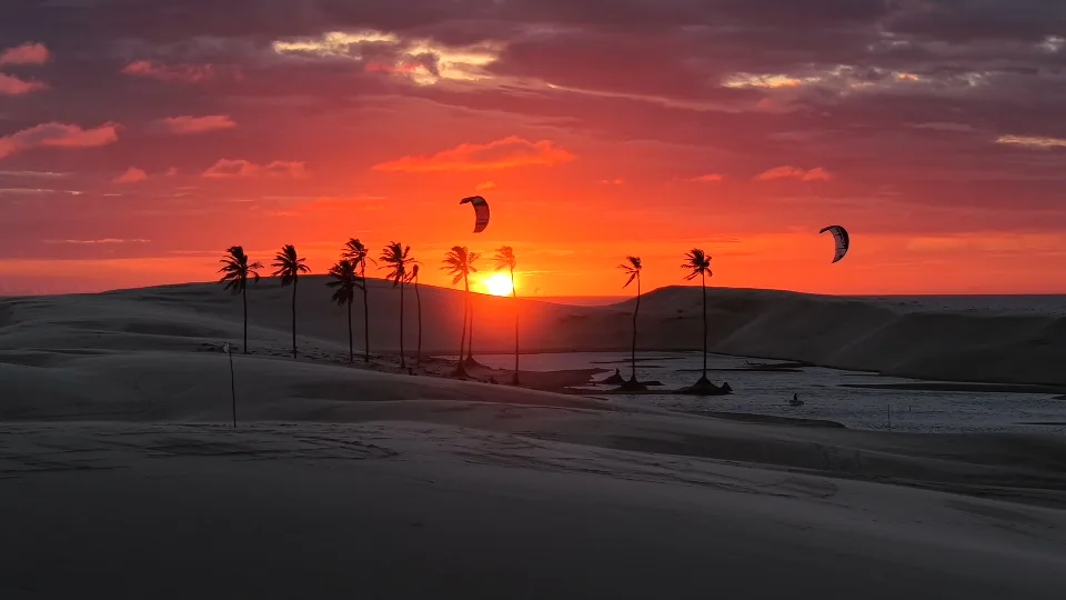 Pôr do sol sobre as dunas e lagoas do Arpoador, com coqueiros e praticantes de kitesurf ao fundo.